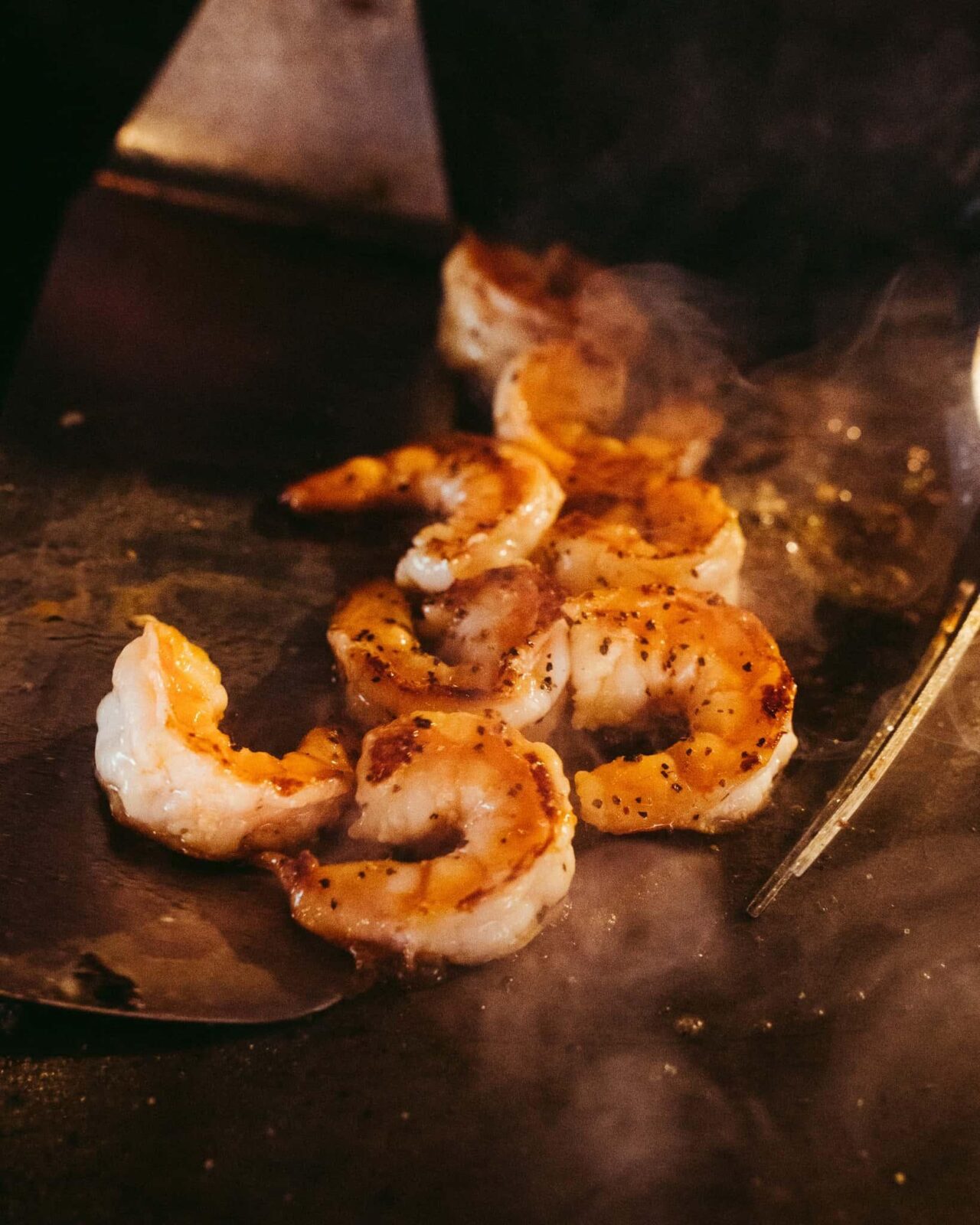 Close-up of several seasoned shrimp cooking on a hot grill with visible steam rising around them, capturing the mouthwatering excitement of a live hibachi dinner—an unforgettable addition to wedding entertainment or a unique Hibachi wedding reception.