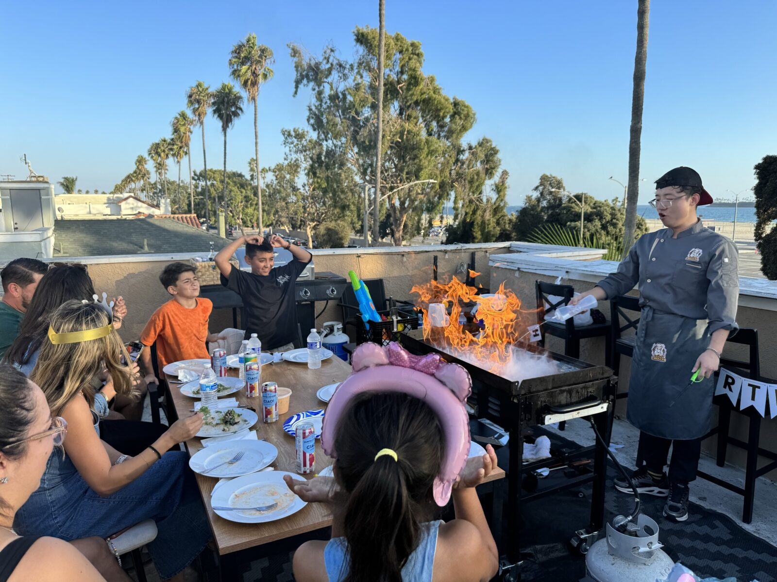 A group of people sit at a rooftop table enjoying a lively hibachi wedding reception, while a chef creates a large flame on the grill. Palm trees and blue sky are visible in the background, adding to the festive wedding entertainment.