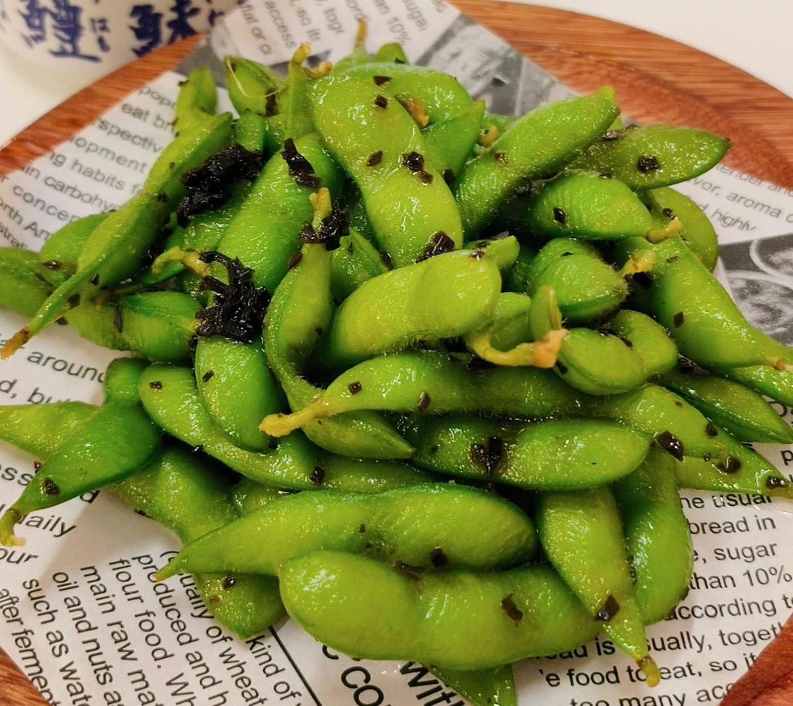 A close-up of cooked edamame pods seasoned with black sesame and spices, served on a piece of printed paper atop a wooden dish—an ideal appetizer for a hibachi wedding reception, where the culinary experience adds to the evening’s unique wedding entertainment.