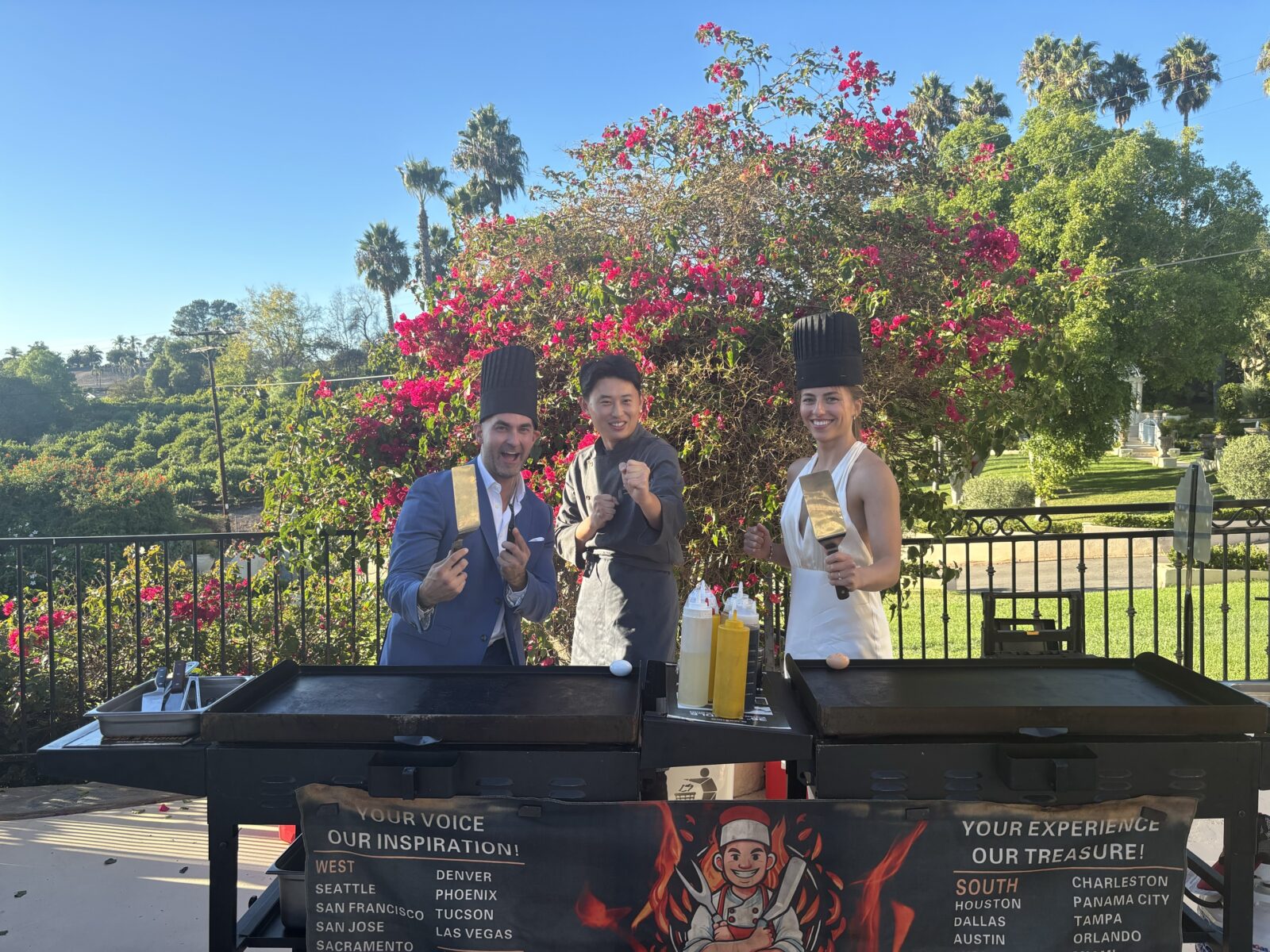 Three people wearing chef hats stand behind outdoor grills, smiling and posing with cooking utensils on a sunny day, with flowers and palm trees in the background, creating the perfect scene for dinner entertainment at a Hibachi wedding.