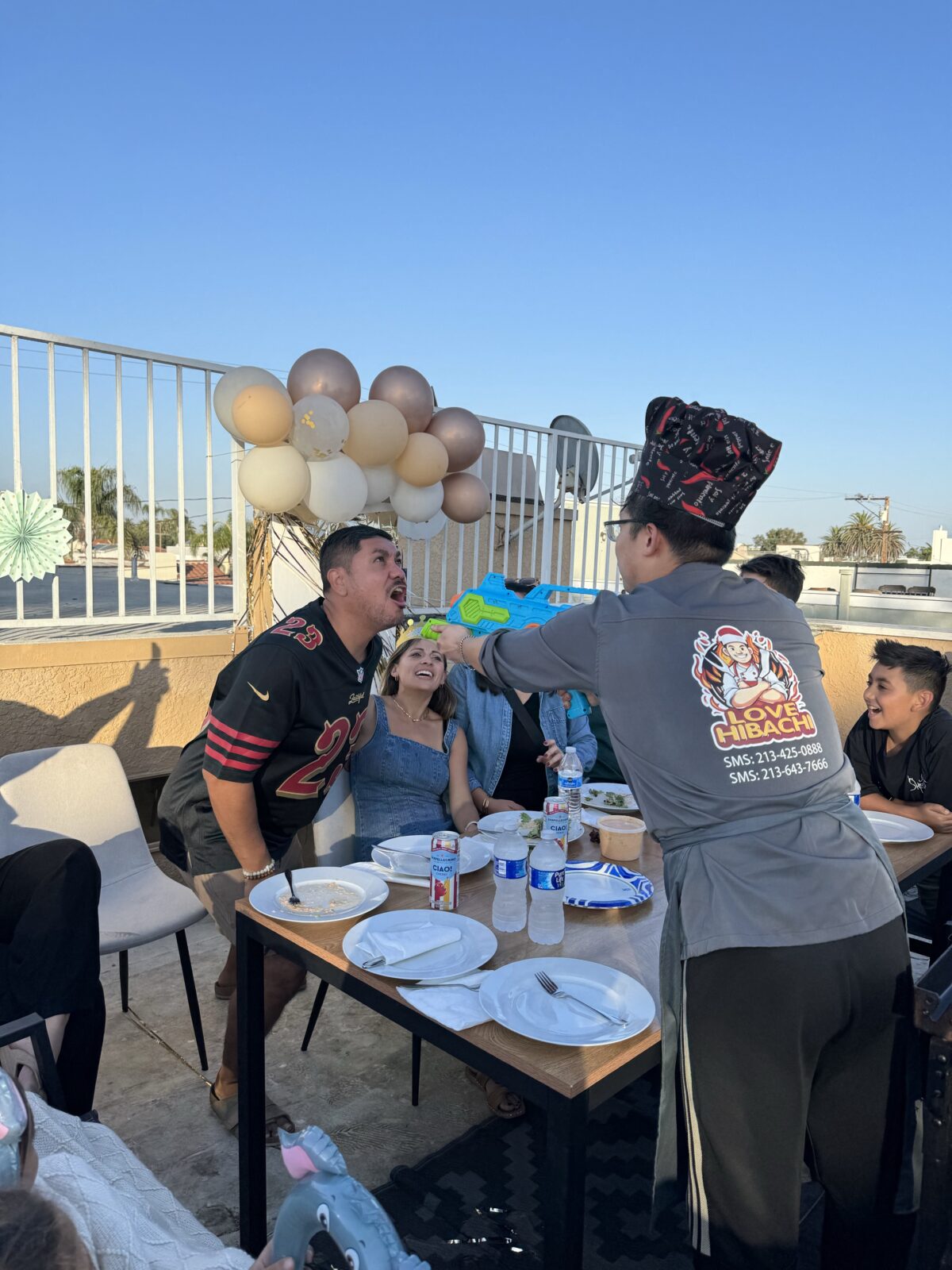 A person playfully sprays another person with a toy water gun at an outdoor gathering, while others watch and laugh, adding a touch of dinner entertainment to the festivities. Balloons and food are on the table, creating a lighthearted atmosphere reminiscent of a lively hibachi wedding reception.