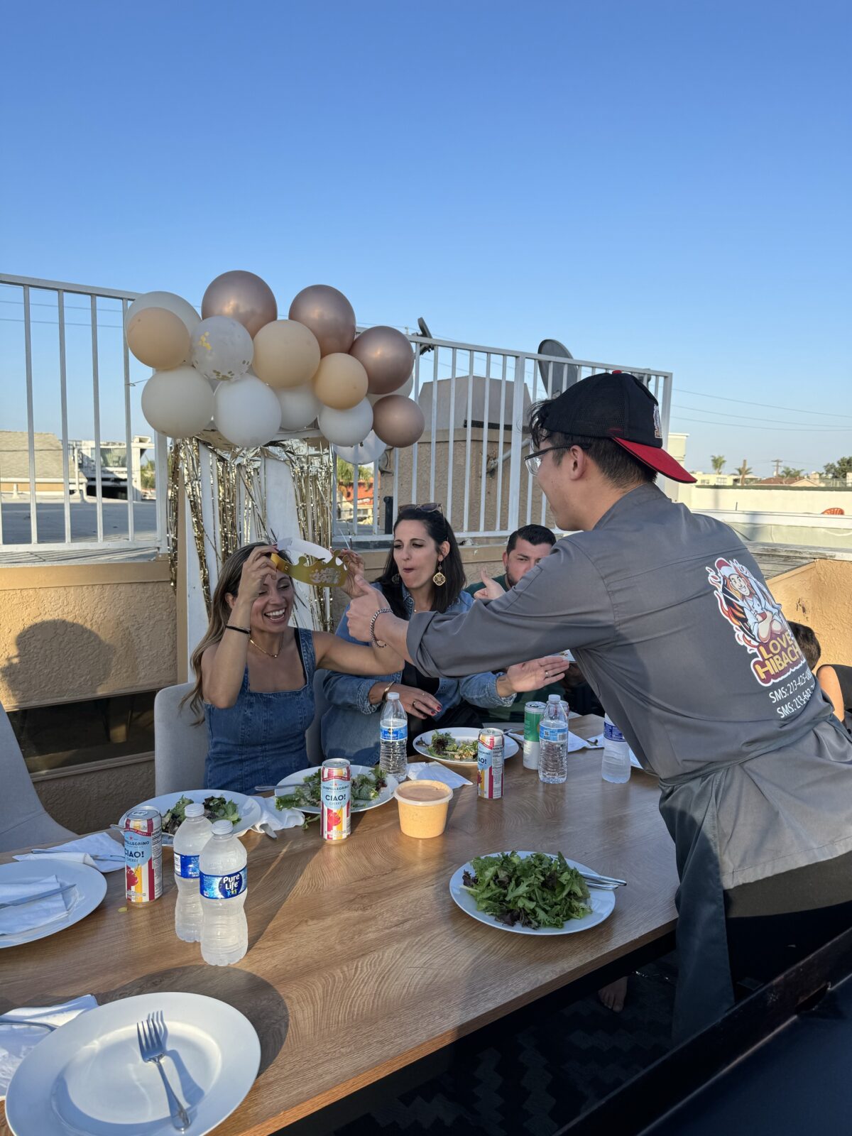 A server hands food to a smiling woman at an outdoor table with plates, drinks, and balloon decorations in the background, creating a lively atmosphere reminiscent of dinner entertainment at a Hibachi wedding.