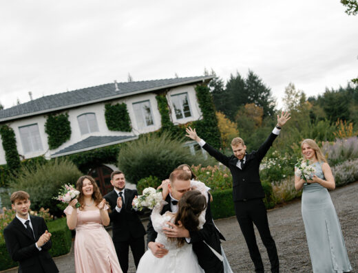 A bride and groom kiss outside a house while their wedding party stands around them, some clapping and smiling, with greenery in the background.