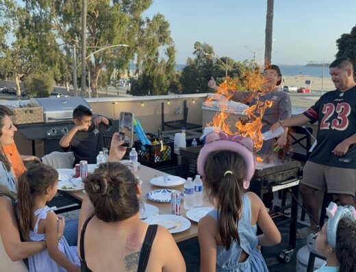 A group of people, including children in party hats, sit around a table outdoors as a grill bursts into flames.