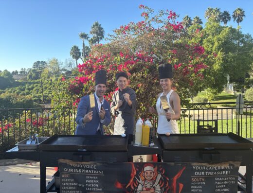 Three people in chef hats stand behind outdoor griddles, posing and smiling with spatulas. A garden with flowers, palm trees, and blue sky is in the background.