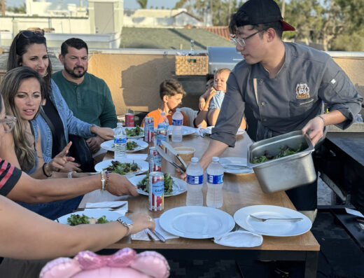 A chef serves salad to a group of people seated at an outdoor table with plates, water bottles, and soda cans on a sunny day.