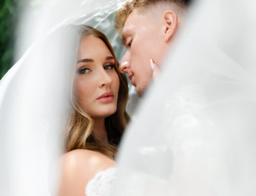 A bride and groom pose closely together under a sheer white veil, with soft natural light and greenery in the background.