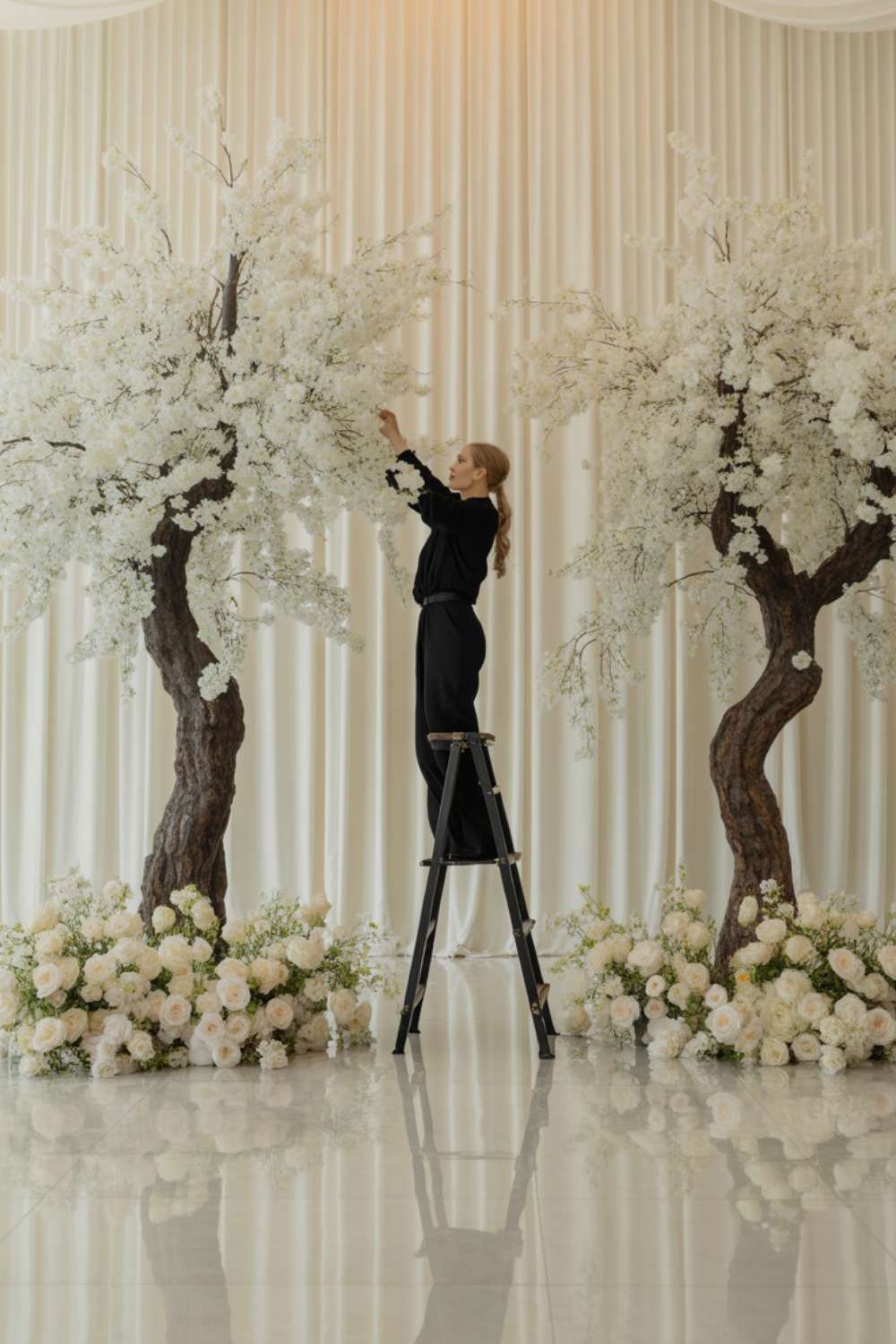 A stylist on a step ladder fluffing the branches of one of two matched white artificial blossom trees against a cream drape wall, with white roses banked around the trunks