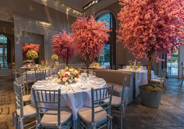 Elegant dining room with round tables set for an event, featuring large pink artificial trees as stunning centerpieces and soft natural light coming through tall windows—perfect inspiration for exquisite wedding decor.