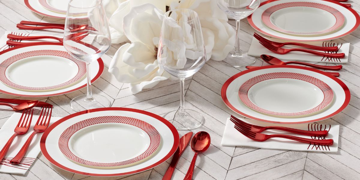 A table set with red and white plates, including elegant disposable wedding plates, red cutlery, wine glasses, and white napkins on a white chevron-patterned surface, with a white floral centerpiece—perfect for stylish wedding table settings.