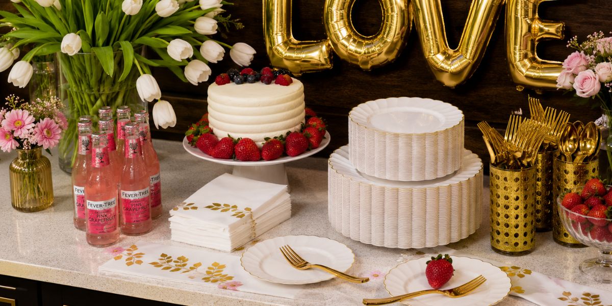 A cake with berries, pink drinks, elegant disposable plates, gold utensils, and flowers are arranged on a countertop with gold “LOVE” balloons in the background—perfect inspiration for chic wedding table settings.