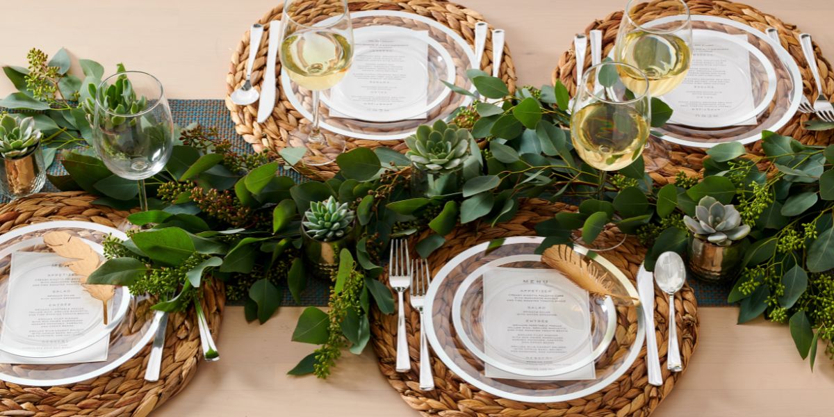 A table set for four with woven placemats, elegant disposable plates, menus, wine glasses, silverware, and green foliage as a centerpiece creates a beautiful wedding table setting.