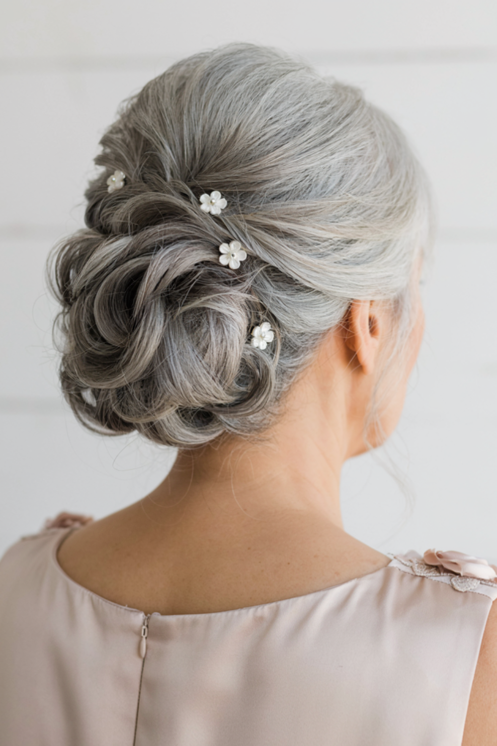 Silver grey curled low chignon with white pearl flower hairpins, shown from the back with a blush dress