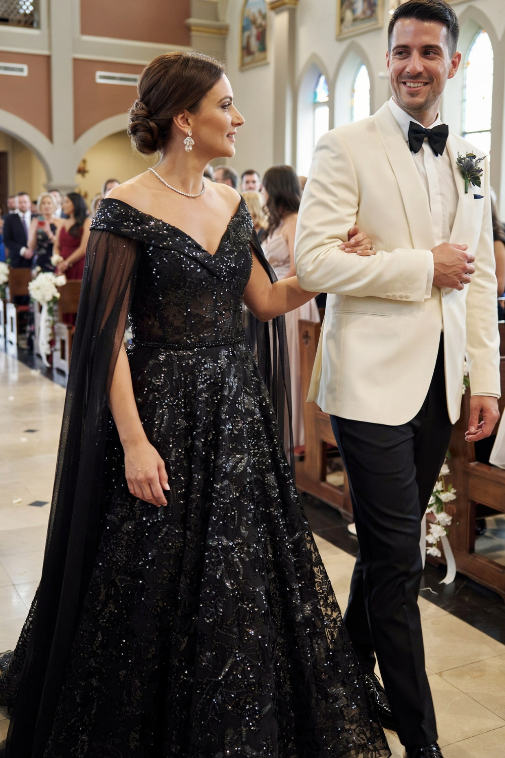 Sleek dark low bun with a deep side part, worn with a black sequin gown and cape while walking down a church aisle