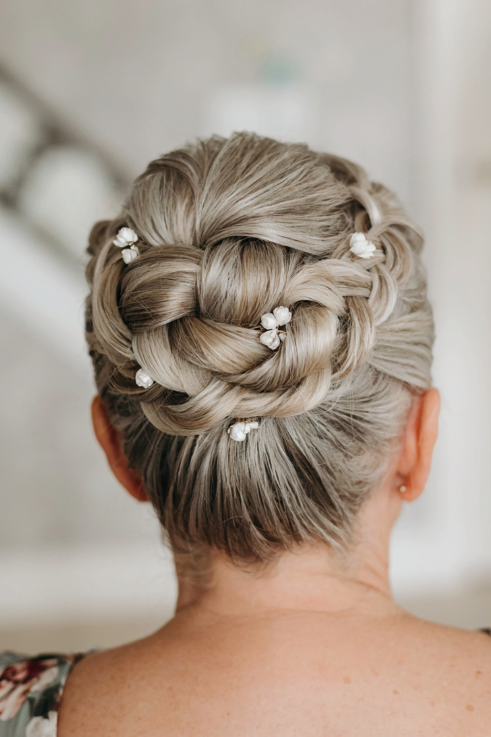 Grey blonde hair braided and coiled into a rosette bun at the crown with small white flower pins