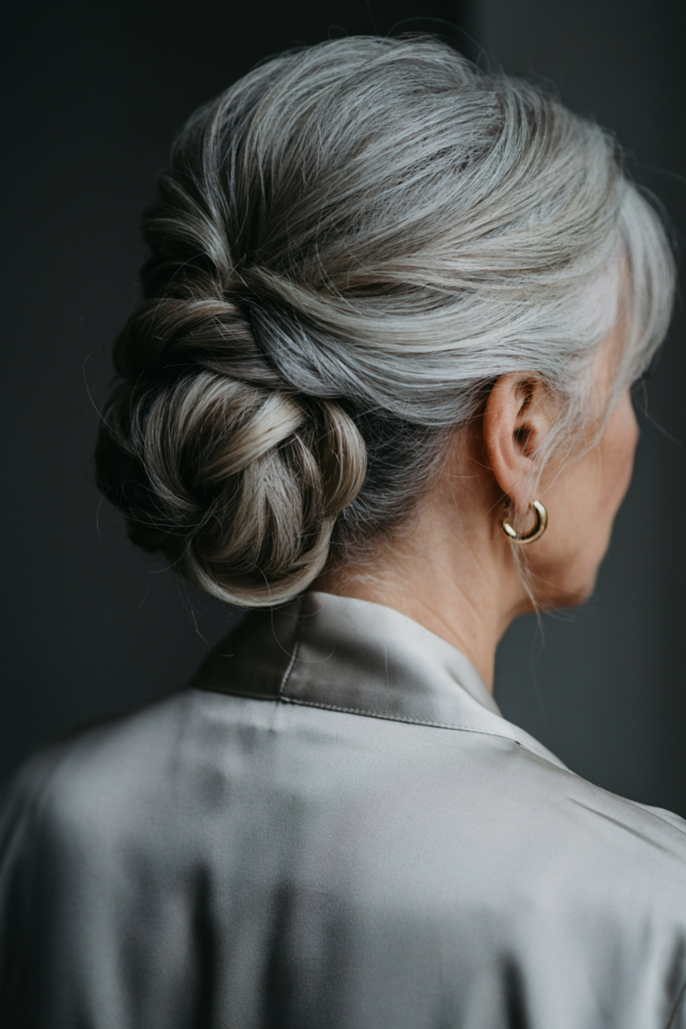 Grey twisted chignon with lifted roots and loose pieces at the temple, worn with a silver satin blouse