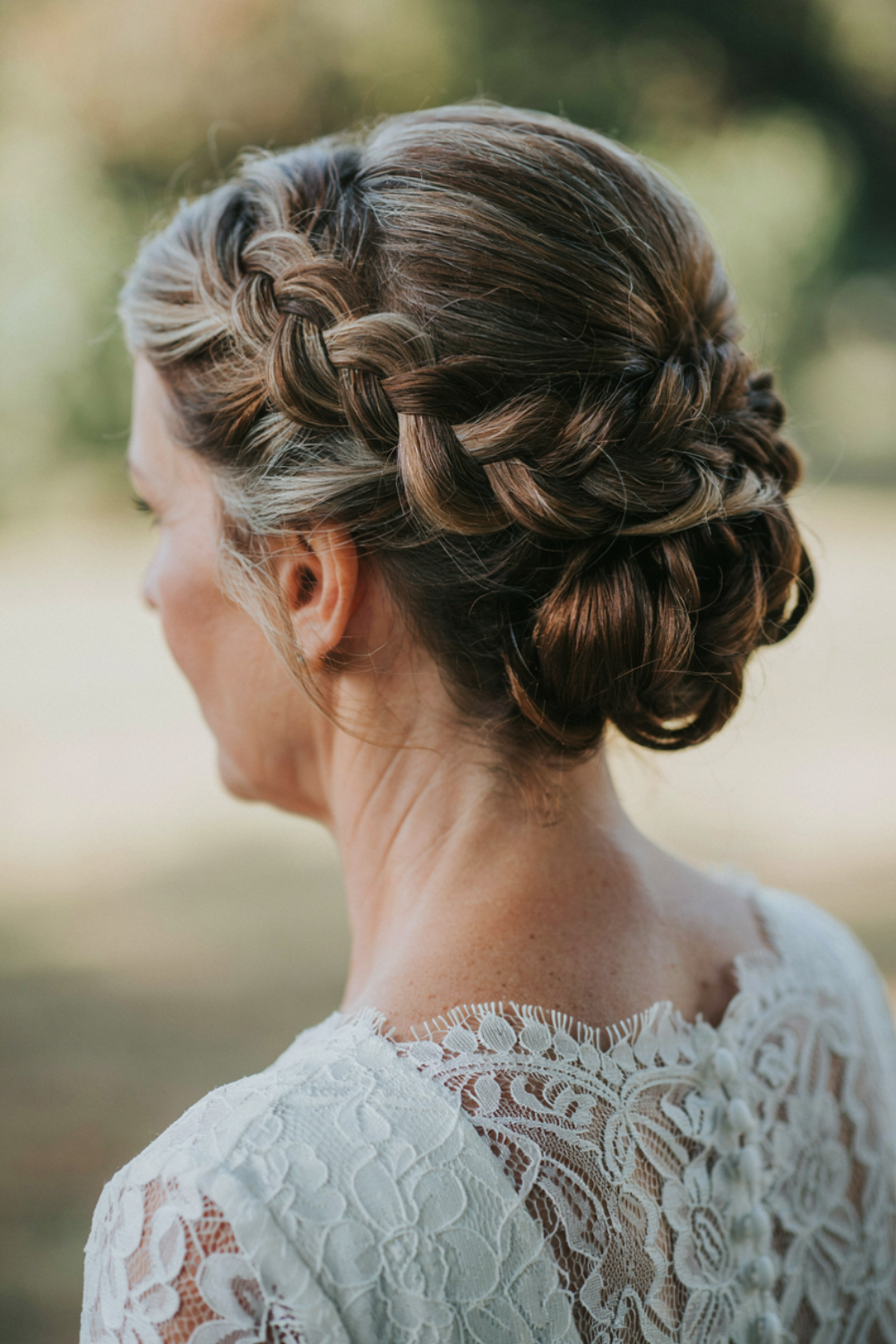 Crown braid across the top of the head leading into a loose textured bun, brunette with highlights, photographed outdoors