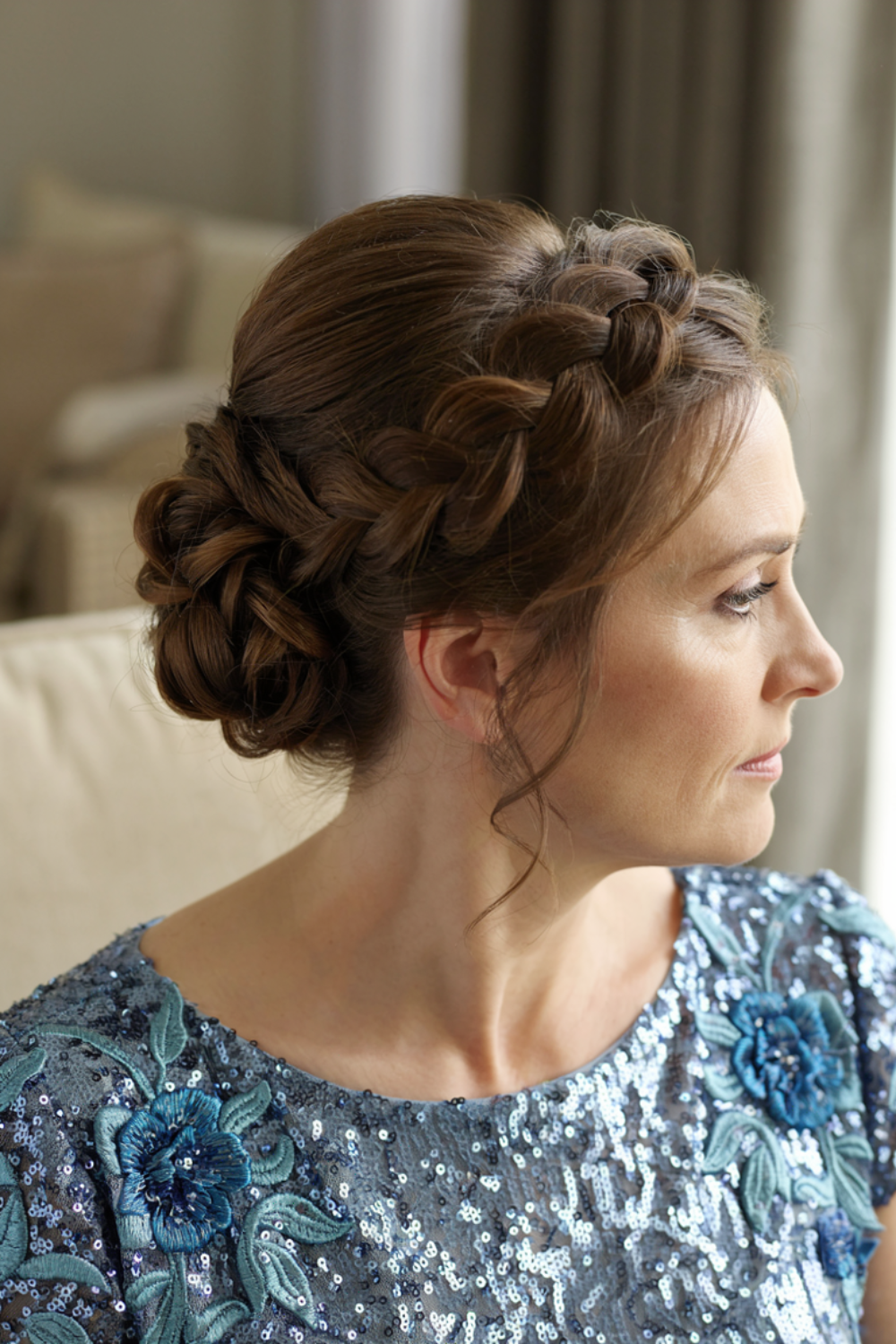 Brunette braided halo leading into a knotted low bun with loose curled tendrils, worn with a blue sequined floral dress