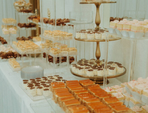 A dessert table with assorted layered pastries and sweets arranged on tiered stands and clear trays, set against a white tablecloth and white walls.