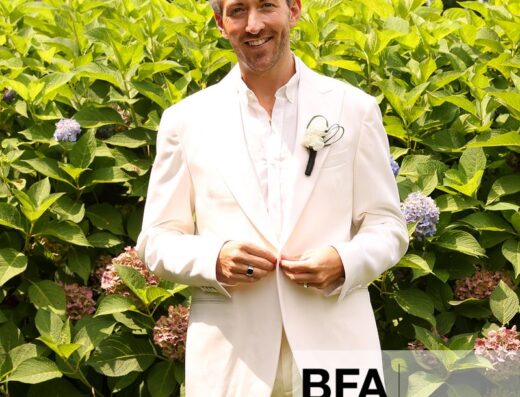 A man in a white suit stands in front of green bushes with hydrangea flowers, smiling at the camera on a beautiful day at a Hamptons Wedding.