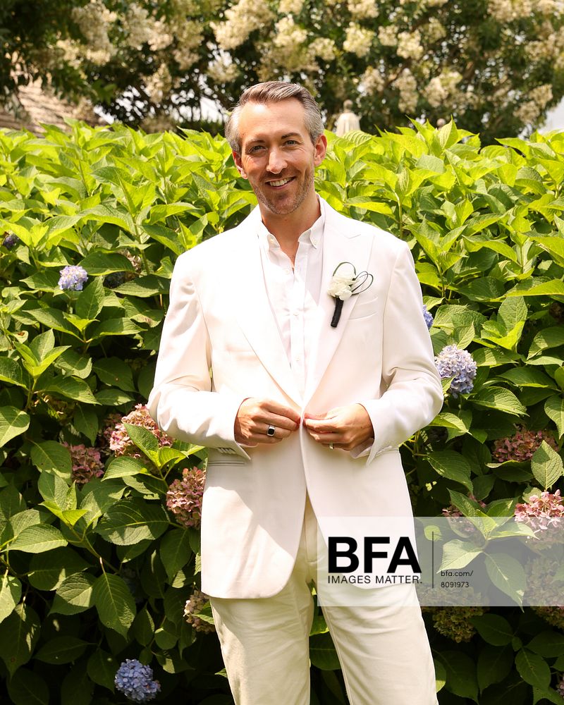 A man in a white suit stands in front of green bushes with hydrangea flowers, smiling at the camera on a beautiful day at a Hamptons Wedding.