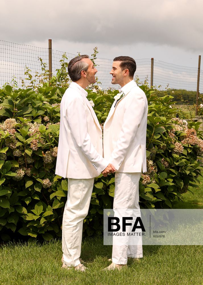 Eric Viner and Jordan Wise, both dressed in elegant white suits, stand face to face, holding hands and smiling outdoors in front of lush green bushes and a wire fence under a cloudy sky—a beautiful moment at their Hamptons Wedding.