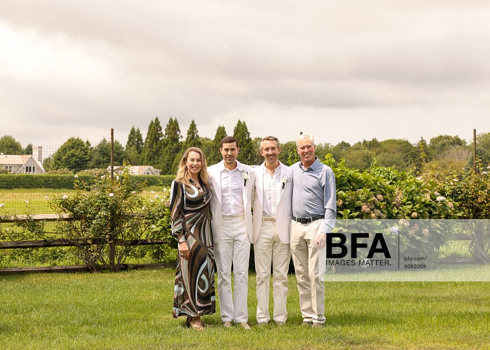 Four adults, including Eric Viner and Jordan Wise, stand side by side on a lawn dressed in formal attire at a Hamptons Wedding, with greenery and houses visible in the background under a cloudy sky.