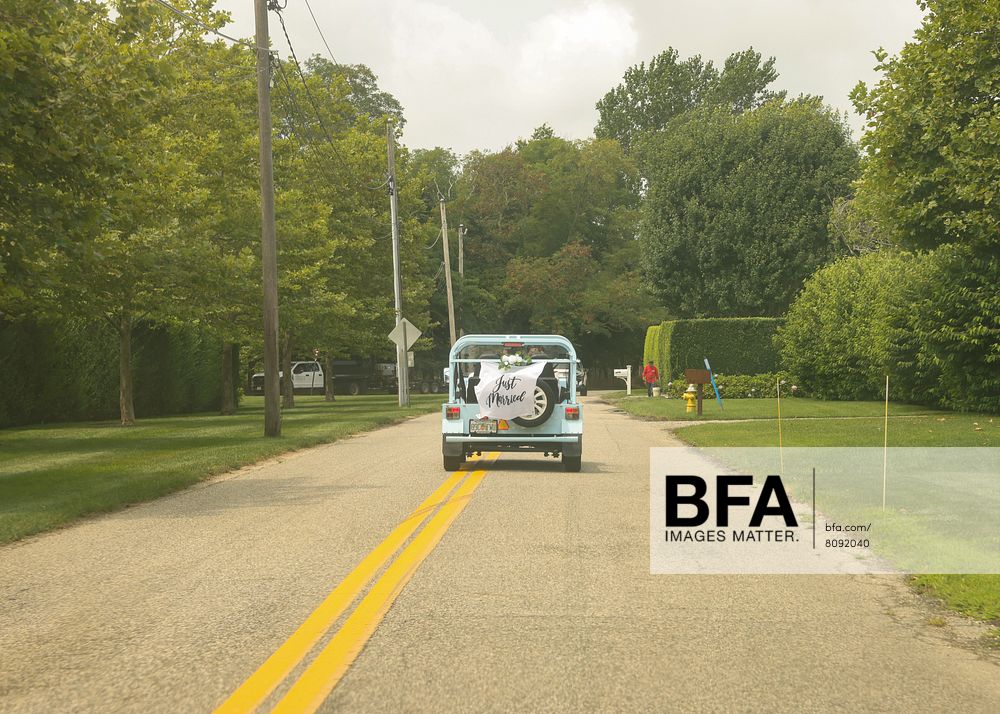 A small blue car with a white “Just Married” sign, fresh from an unforgettable Hamptons Wedding, drives down a suburban tree-lined street on a cloudy day.