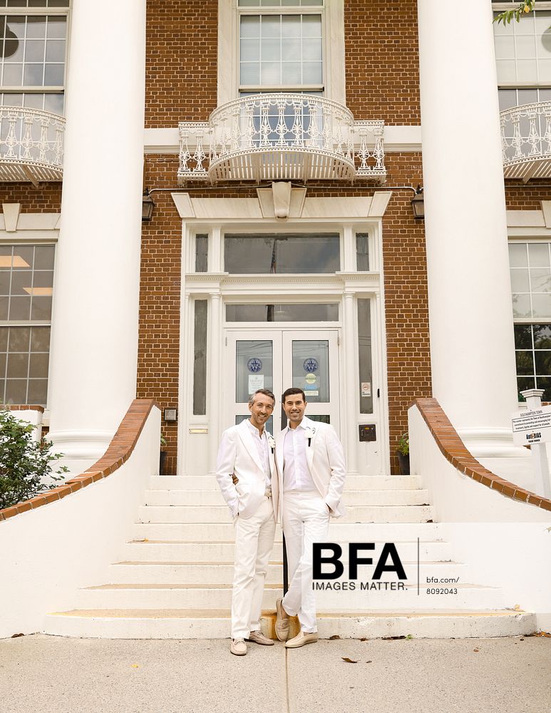 Two men in white suits, Jordan Wise and Eric Viner, stand together on the steps of a brick building with columns and a balcony, posing for a photo at their elegant Hamptons wedding.