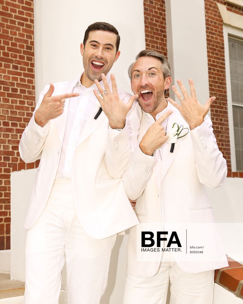 Two men in white suits stand outside, smiling and pointing to rings on their left hands, suggesting they have just gotten married at a beautiful Hamptons wedding.