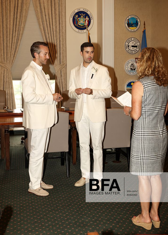 Eric Viner and Jordan Wise, both dressed in white suits, stand facing each other holding hands as a woman holding papers officiates their Hamptons wedding in a formal room adorned with official seals on the wall.