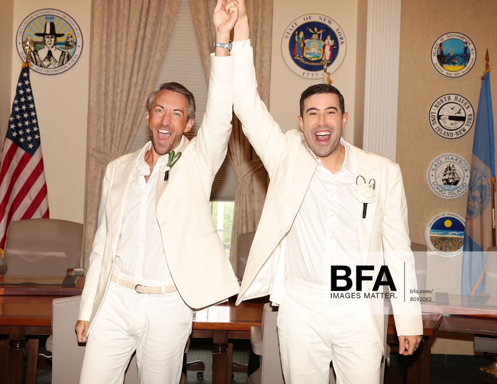 Two men in white suits, Eric Viner and Jordan Wise, stand in a government office, smiling and raising their joined hands in celebration. Official seals and flags are visible in the background, marking the joyous occasion reminiscent of a Hamptons Wedding.