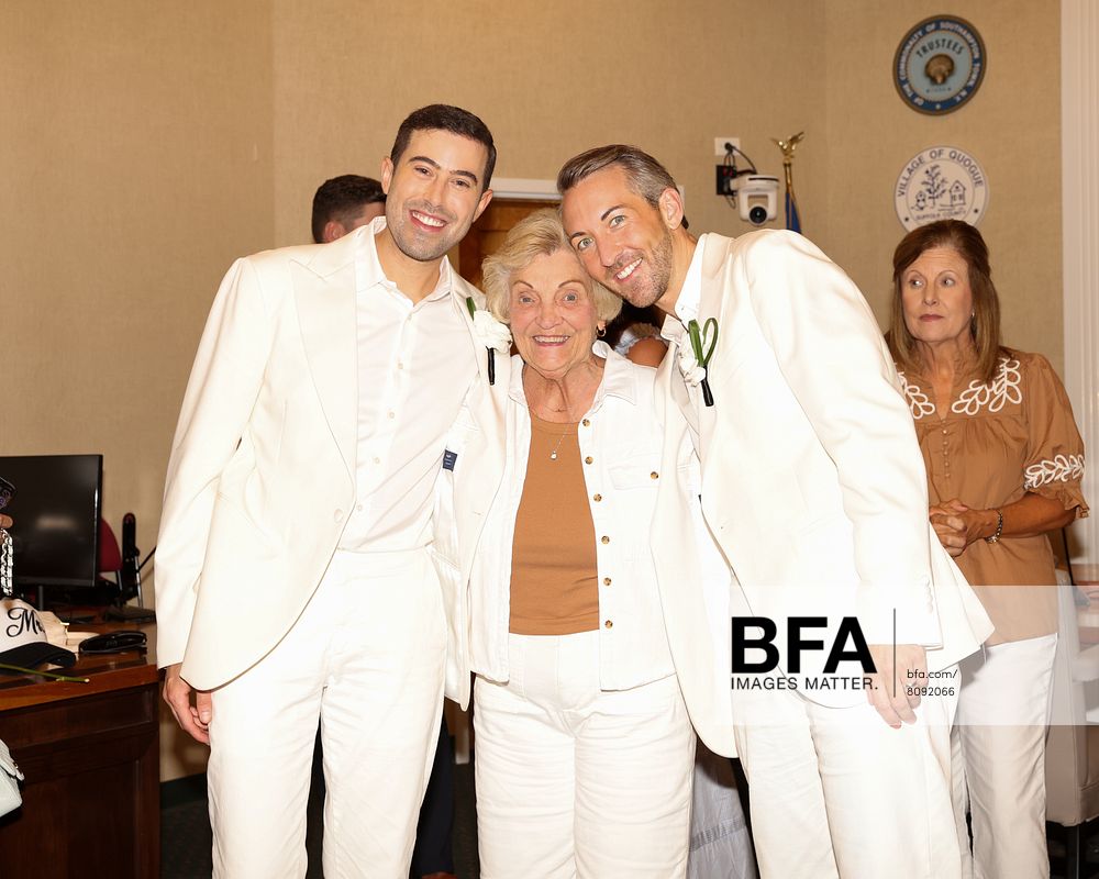 Three people in white suits pose and smile together indoors; two men—one identified as Eric Viner—stand on either side of an older woman. A woman in the background watches. A “STATE OF GEORGIA” seal is on the wall, suggesting a formal occasion reminiscent of a Hamptons Wedding.