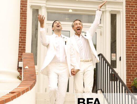 Two men in white suits, Jordan Wise and Eric Viner, hold hands and celebrate on the steps outside a building, smiling as flower petals fall around them—a joyful moment reminiscent of a Hamptons wedding.