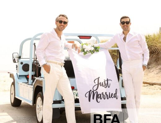 Two men in white outfits, Jordan Wise and Eric Viner, stand by a blue car with a “Just Married” sign on the back, smiling on a sunny day after their Hamptons Wedding.
