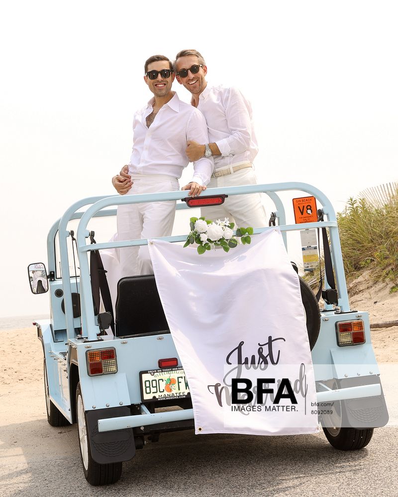 Two men in white shirts and pants stand on the back of a blue convertible with a “Just Married” sign and flowers, parked near a beach—capturing the joyful spirit of Eric Viner and Jordan Wise’s Hamptons wedding celebration.