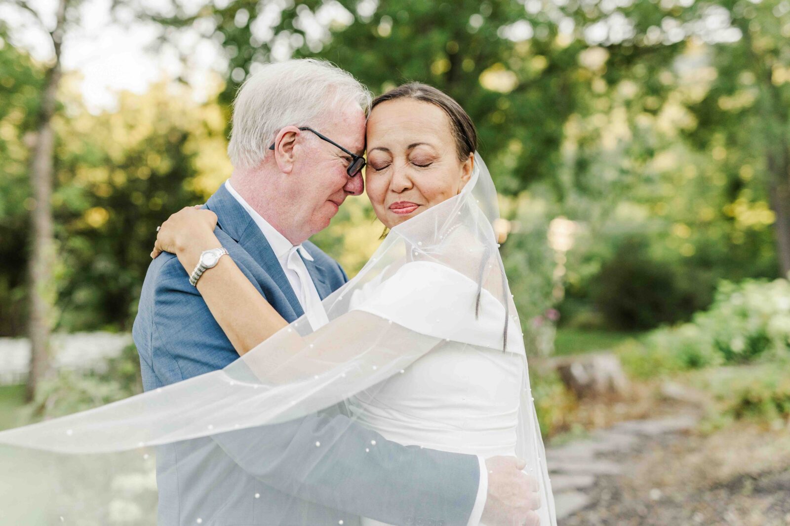 An older couple embraces outdoors, the woman in a white dress and veil and the man in a blue suit, with greenery in the background.