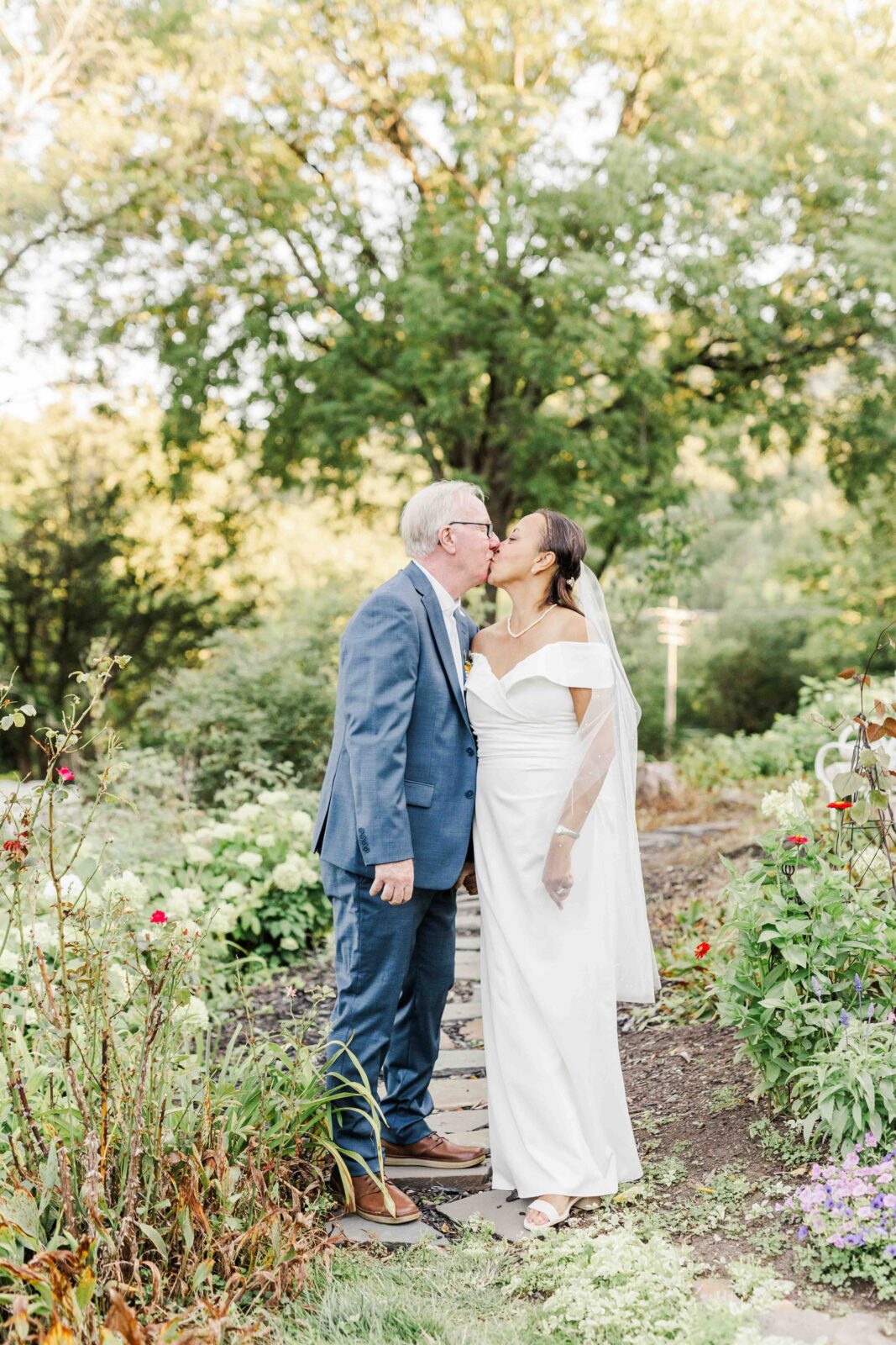 A couple dressed in wedding attire shares a kiss on a garden path, surrounded by greenery and flowers, with trees in the background.