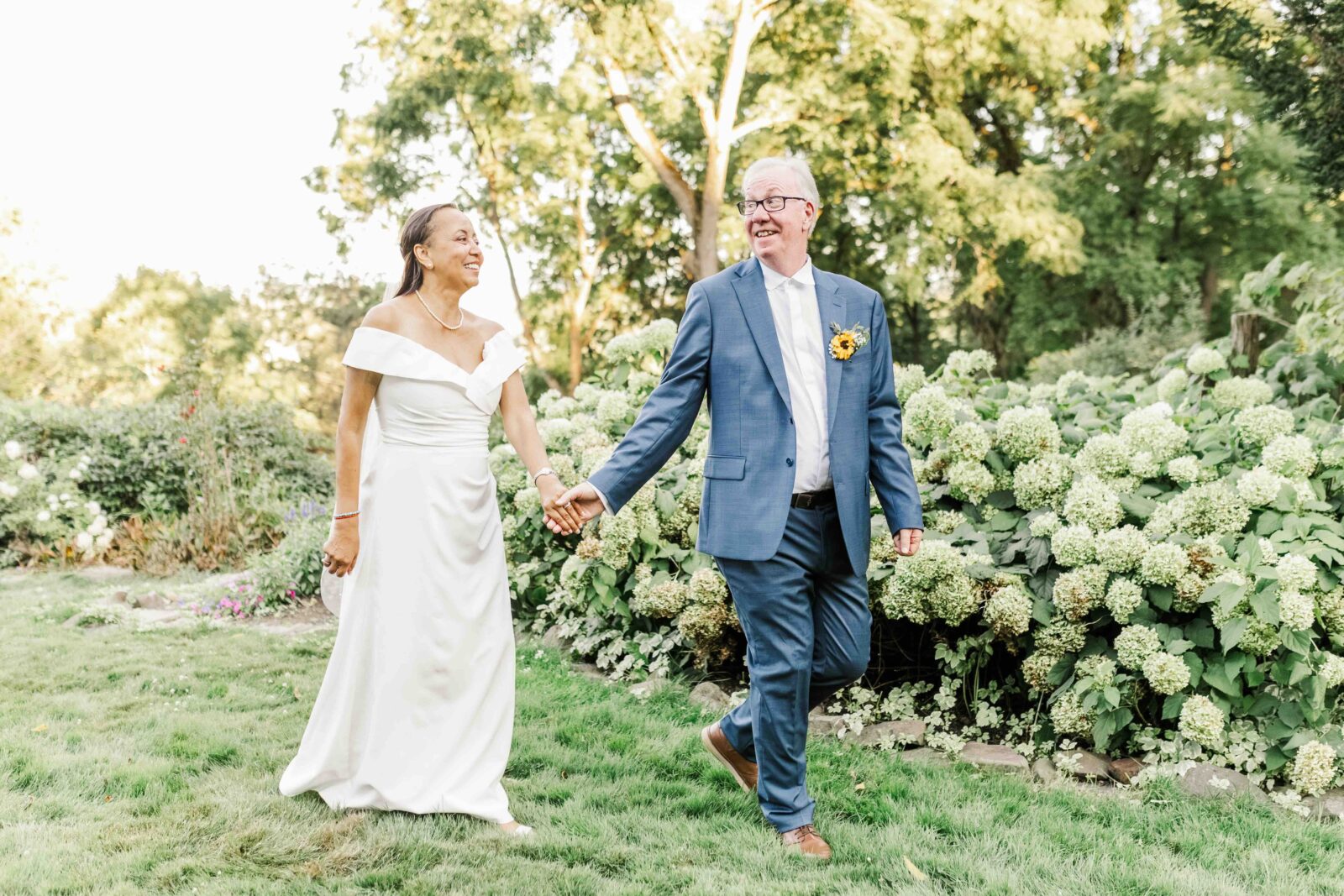 A couple, dressed in wedding attire, walks hand in hand on a grassy path surrounded by green bushes and trees on a sunny day.