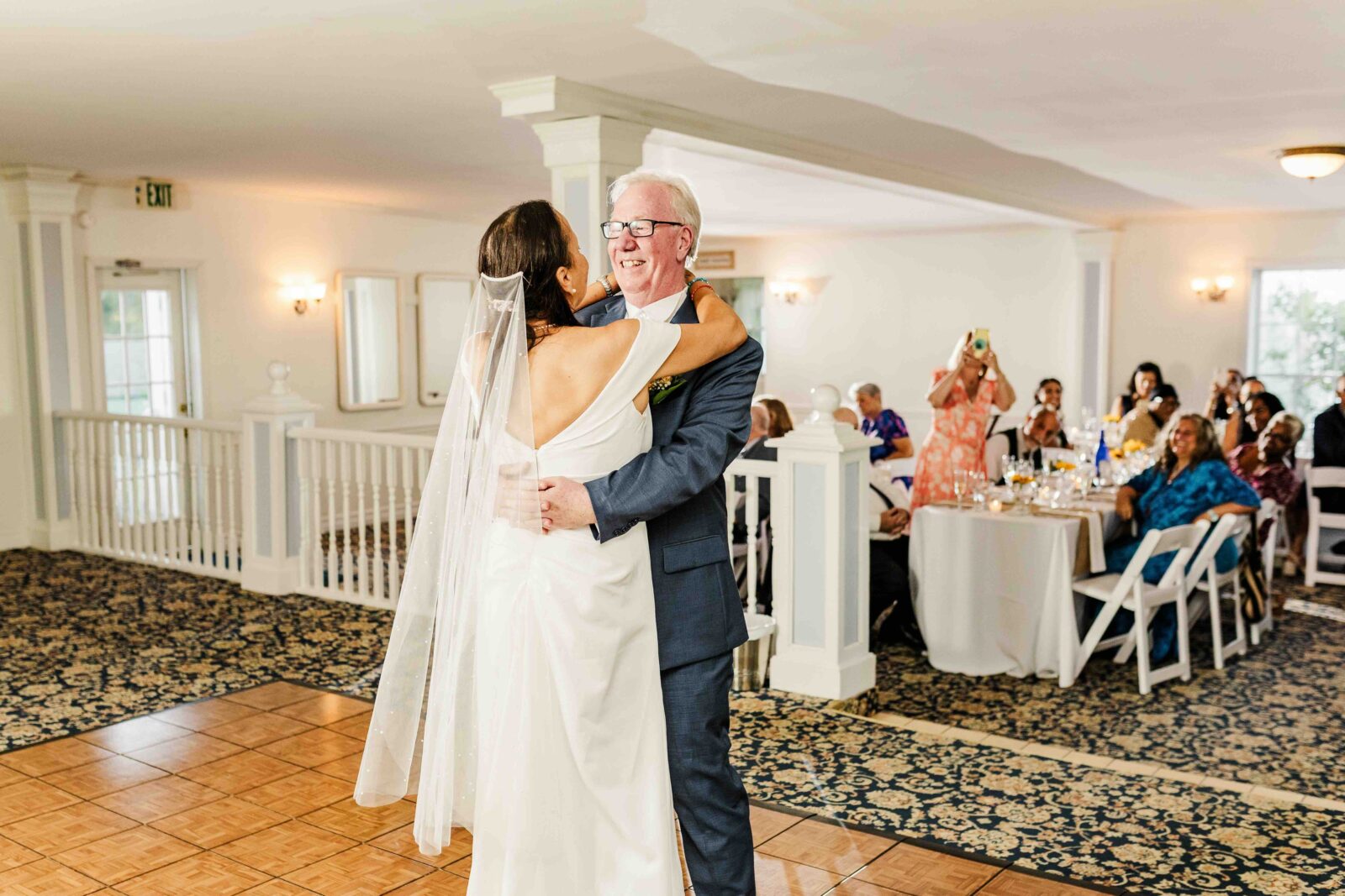 A bride in a white gown and veil dances with an older man in a blue suit at a wedding reception while guests watch from tables in the background.