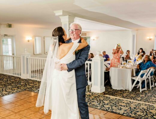 A bride in a white gown and veil dances with an older man in a blue suit at a wedding reception while guests watch from tables in the background.
