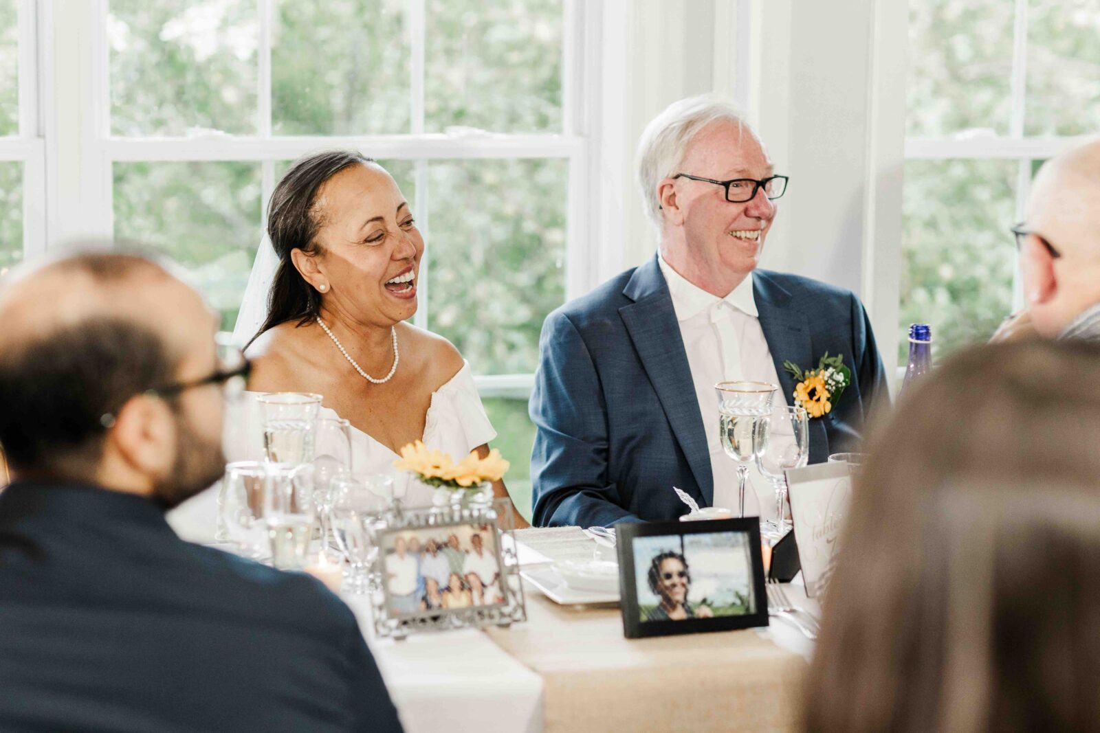 A smiling couple sits at a decorated table during a celebration, surrounded by guests, glasses, framed photos, and flowers.