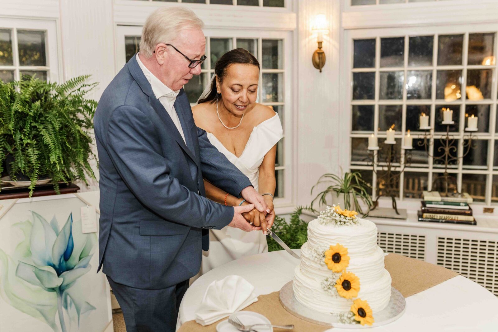 A couple in formal attire stands together, smiling as they cut a white, tiered cake decorated with sunflowers in a warmly lit room.