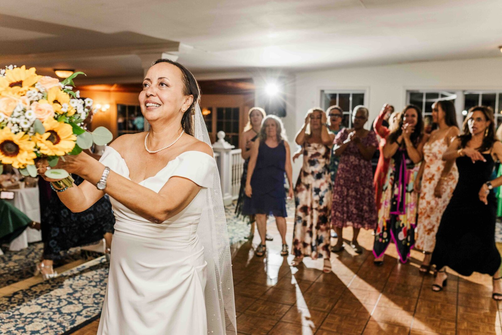 A bride in a white dress holds a bouquet, preparing to toss it to a group of women standing behind her at a wedding reception.