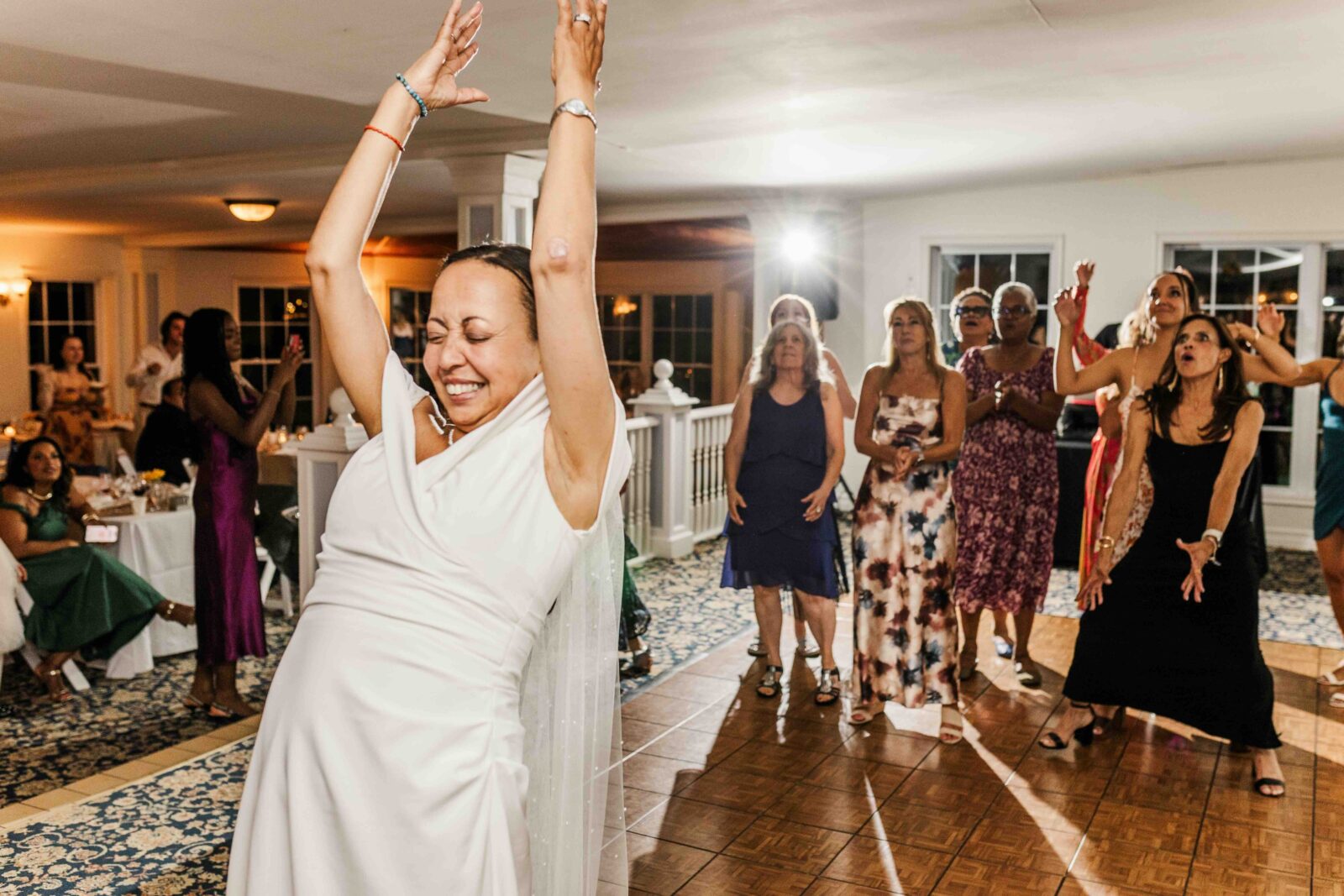 A bride with arms raised stands in front as a group of women eagerly reach out behind her in a brightly lit wedding reception hall.
