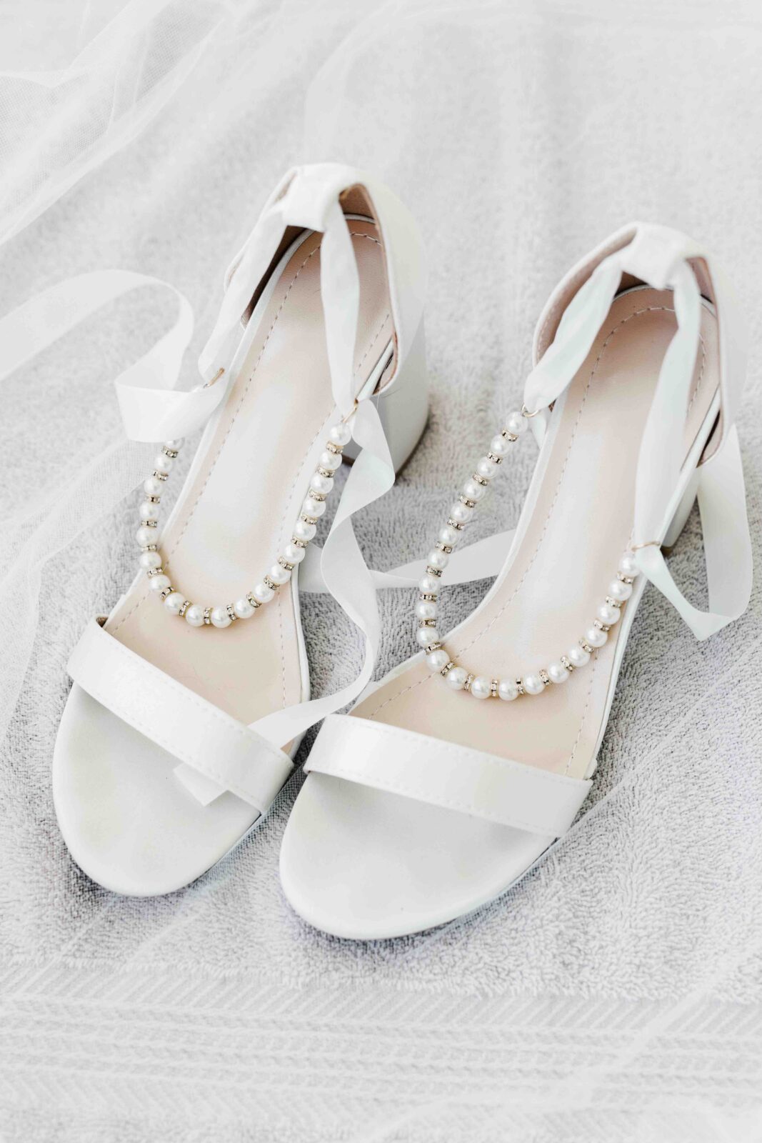 A pair of white open-toe high heel shoes with ankle ribbons and pearl decorations, placed on a light textured surface.