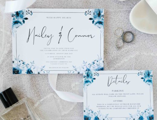 Flat lay of two floral wedding invitations, perfume bottle, two rings, white sandals, and blue flowers on a textured light surface.