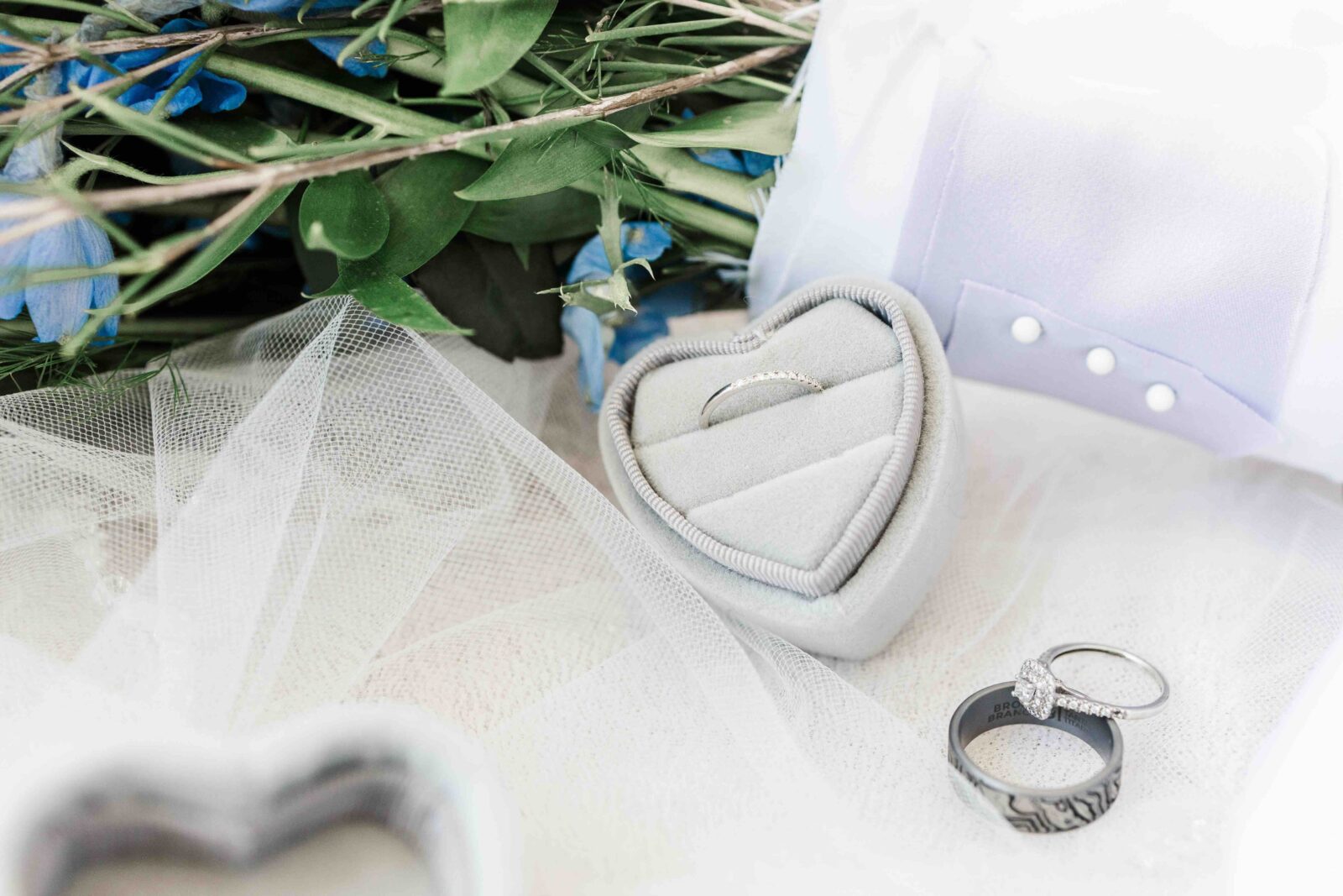 Two rings in a heart-shaped box, one ring and a wedding band nearby, with a bouquet of blue flowers and sheer white fabric in the background.