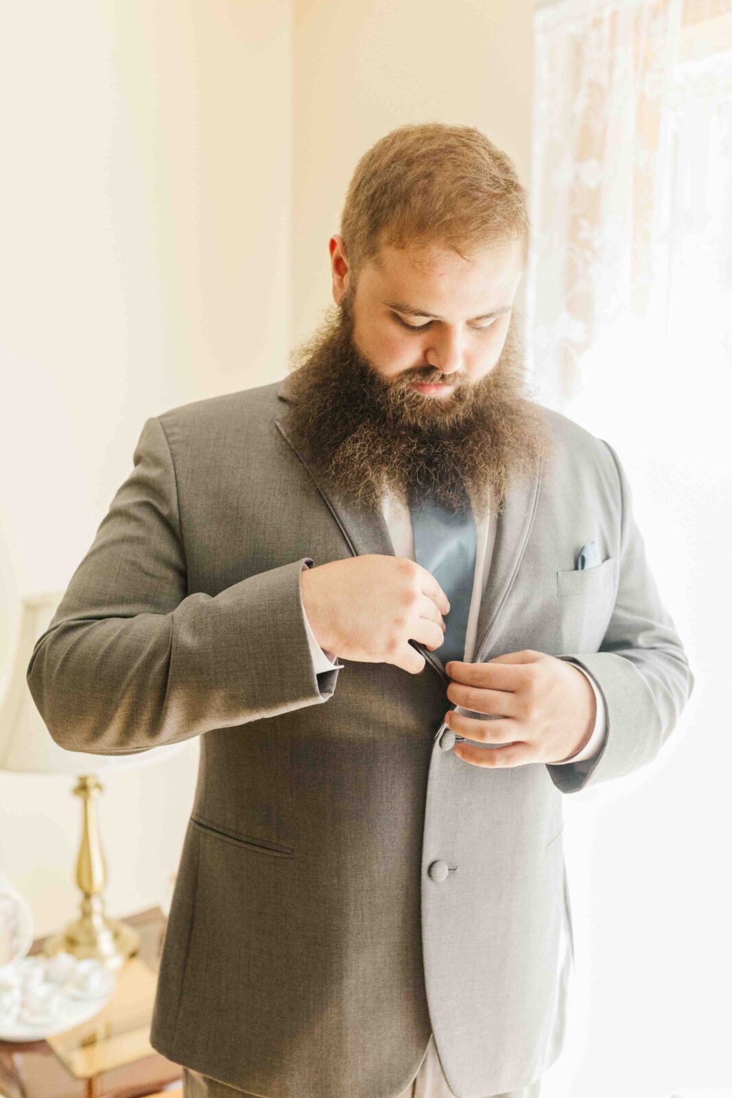 Man with a long beard wearing a light gray suit is buttoning his jacket in a well-lit room near a window with lace curtains.