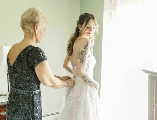 A woman helps a bride in a white gown with floral details adjust her dress near a window in a softly lit room.